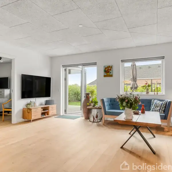 Stue med blå sofaer og et sofabord i midten, fladskærms-tv monteret på væggen. Store vinduer med udsigt til have, træmøbler og dekorationer skaber en lys og moderne atmosfære.