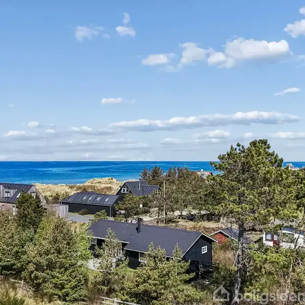 Træhuse står stille blandt grønne træer nær en kyst med blåt hav og blå, let overskyet himmel i baggrunden.