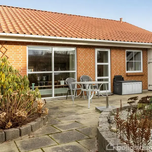 Et lille bord med to stole står på en flisebelagt terrasse foran en rød murstensbygning med rødt tegltag. Omgivet af planter og en lille stenhave.