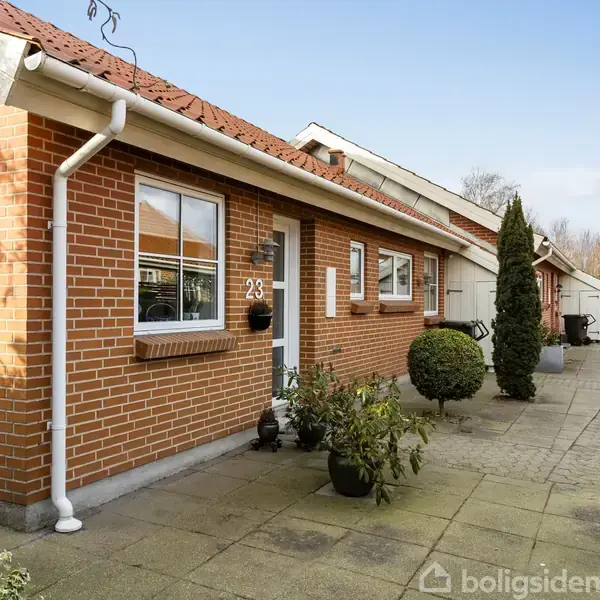 Rødt murstenshus med nummeret 23 ved hoveddøren. Huset har en asfalteret indkørsel med nogle planter og buske omkring. Skyfri himmel i baggrunden.