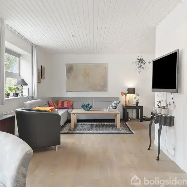 Stue med sofagruppe rundt et sofabord, dekoreret med puder. Tv hænger på væggen. Store vinduer giver naturligt lys. En blød lænestol står foran et sidebord med lyseholder.