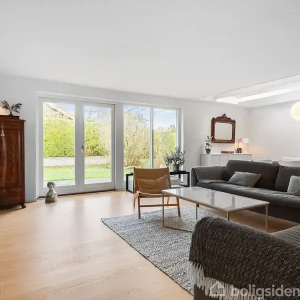 TV står på et skab i en lys stue med en stor grå sofa og udsigt til en have gennem glasdøre. Væggene har kunstværker, og et loftlys hænger over sofabordet.