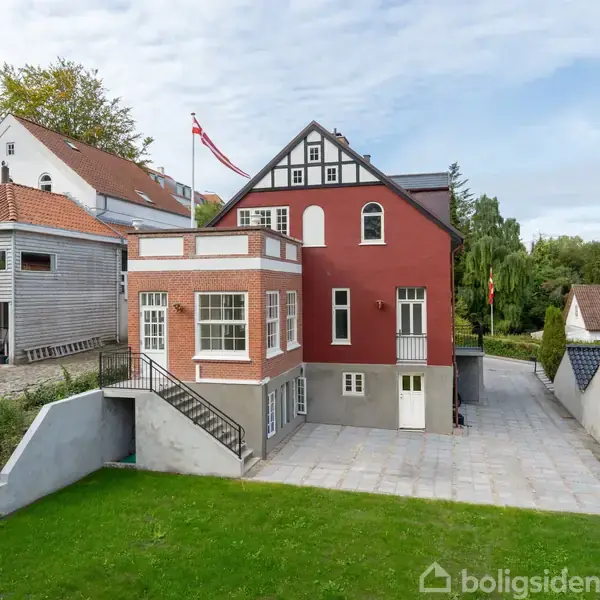 Et stort, rødt murstenshus med hvide vinduer og sort-taget gavl, der står på en flisebelagt grund. I baggrunden er grønne træer og et gråt træhus med rødt tegltag.