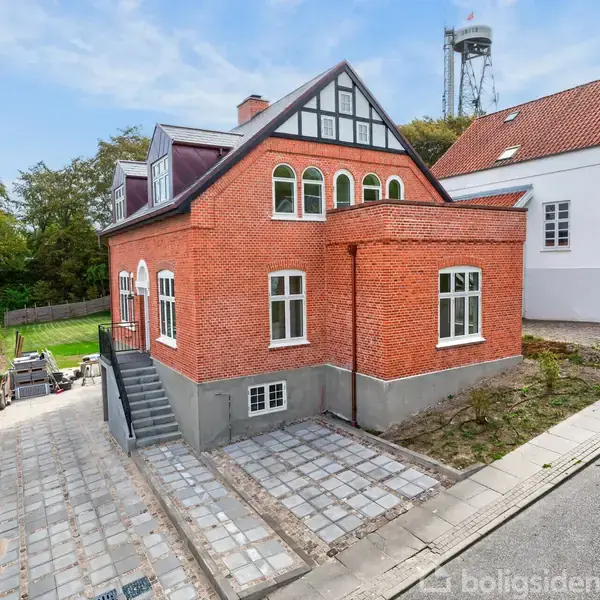 Rødt murstenshus står stille på en brolagt indkørsel i et villakvarter. Træer ses i baggrunden, mens en hvid bygning er tættere på, til højre.