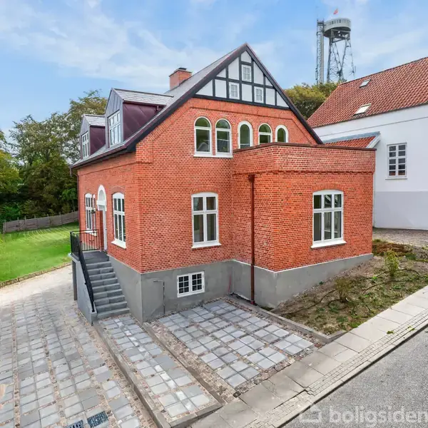 Rødt murstenshus står foran skovklædt område, flankeret af indkørsel med fliser. Hvidt hus ses til højre, og en vandtank i baggrunden.