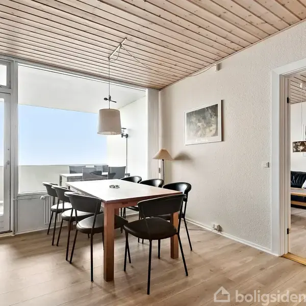 Spisebord med sorte stole står i en spisestue. Der hænger en lampe over bordet, og et vindue åbner ud til en balkon. Til højre ses en stue med sofa.