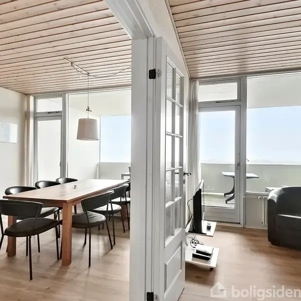 Spisebord med sorte stole står i en lys stue med trægulv og træloft. Store vinduer og glasdør åbner mod en altan. Skænk og lænestol i rummet.