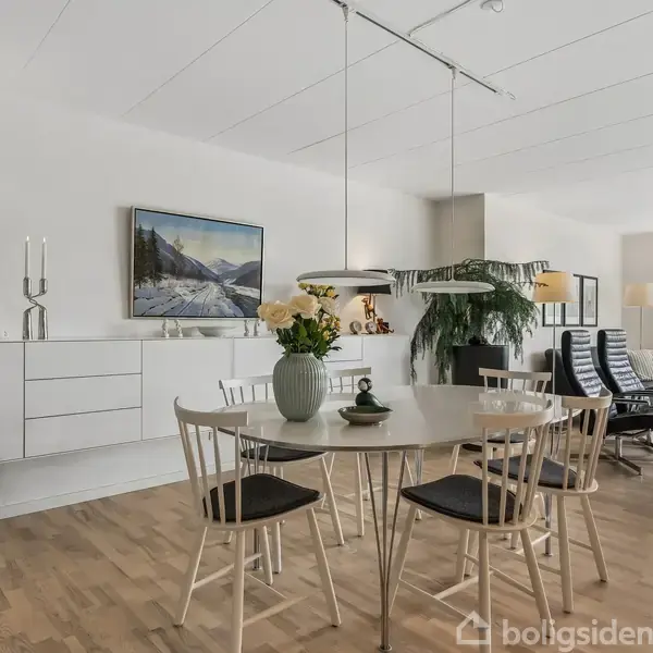 Et lyst spiseområde med et rundt bord og seks stole. En vase med blomster står på bordet. I baggrunden ses en moderne stue med lænestole og dekorationer.