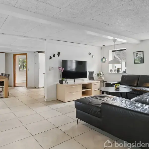 Sort lædersofa står i lyst, rummeligt stueområde med flisegulv. Foran er et tv på træmøbel. Spisebord med stole til venstre ved et vindue. Rummets vægge er hvide.