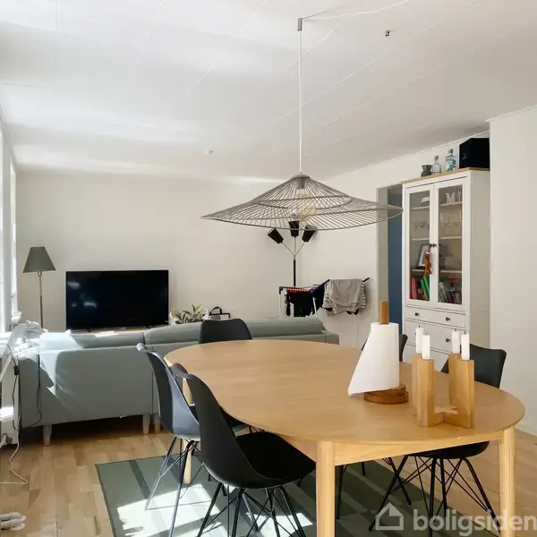 Spisebord omgivet af sorte stole står under en stor lampe. I baggrunden ses en sofa ved siden af et tv. Rummet er lyst med hvide vægge og trægulv.