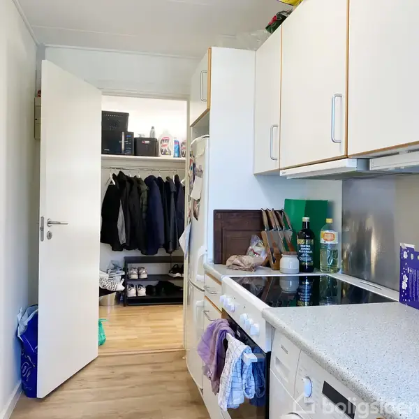 Køkkenet har en hvid bordplade med kogeplader og forskellige køkkenartikler. En åben dør fører til et rum med tøj hængende på en bøjlestang og hylder med kasser og rengøringsmidler.