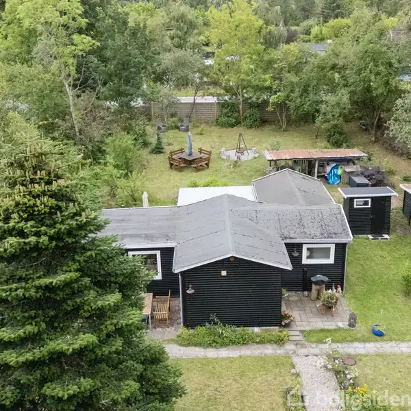 Et sort malet sommerhus med gråt tag står i en grøn have omringet af høje træer. En grussti leder til hoveddøren. En carport ses til højre.