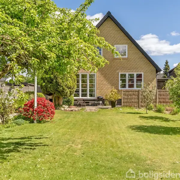 Et gulstenshus med trekantet tag står i en have med grønne græsplæner og blomstrende buske. Der er blå himmel og få, hvide skyer i baggrunden.