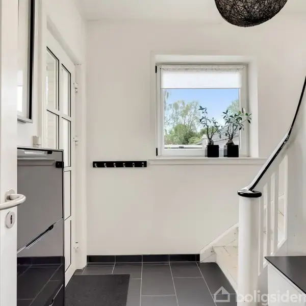 En lys entré har en hvid trappe til højre, der fører opad. Et vindue med grønne planter står bagved. Døren til venstre er flankeret af en sort kommode.