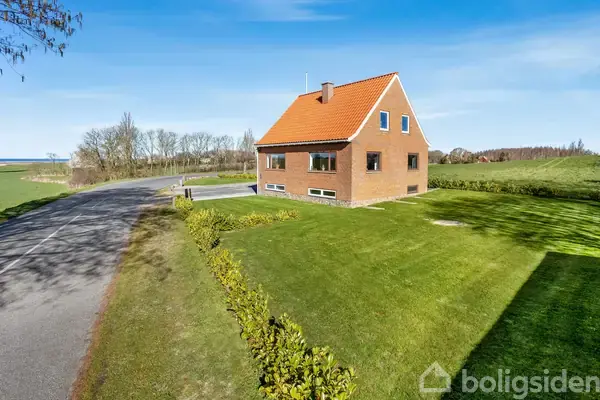 En rød murstensbygning står ved en græsplæne. En bred vej ses i baggrunden med åbne marker og spredte træer, hvilket giver en landlig atmosfære.