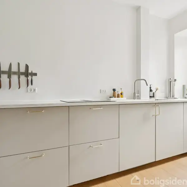 Et moderne køkken med lysegrå skabe, knive hængende på væggen, vask og diverse køkkenartikler. Store vinduer lader naturligt lys ind, fremhæver rummets minimalistiske stil.