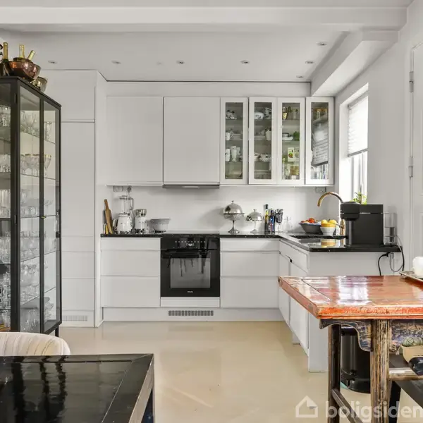 Et hvidt køkken med moderne skabe og glaslåger, hvor der står køkkenredskaber og frugtskål. Ved siden af er en glasvitrine med glas, og der er et spisebord med lysestager.