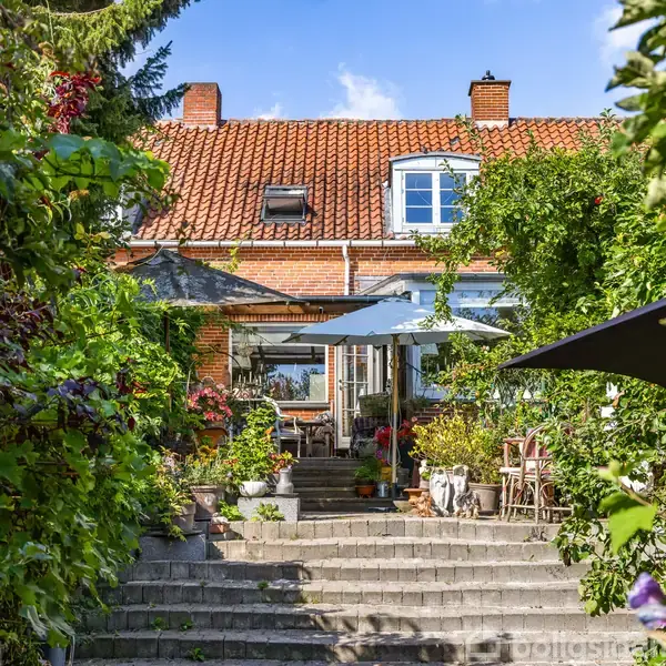 En charmerende murstensvilla med rødt tegltag og hvide vinduer, omkranset af frodige planter og trapper, leder hen til en terrasse med parasoller og havemøbler i solskinnet.