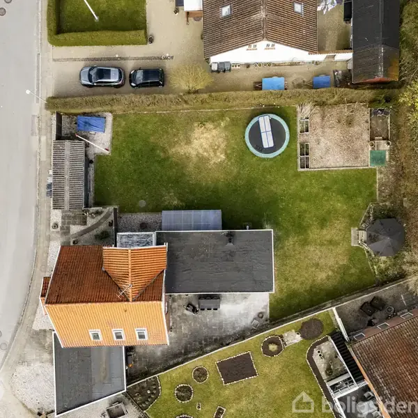 Et hus set ovenfra har en rød tag og en større have med en trampolin. Flere biler er parkeret langs en vej til venstre. Omkringliggende boliger omgiver ejendommen.