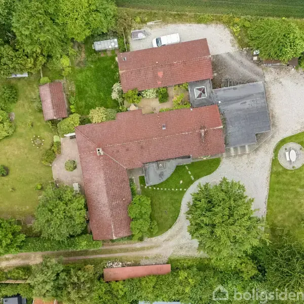 Stor gård med røde tage omgivet af omfattende grønne områder og haver. En grusvej fører til bygningen, hvor to biler er parkeret tæt på. Træer omkranser ejendommen.