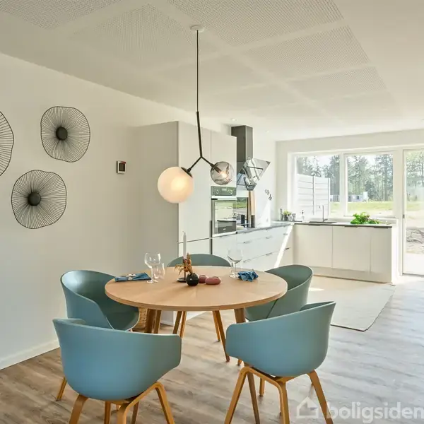 Rundt spisebord med fire blå stole står i et lyst, åbent køkken-alrum. Dekorative vægskiver hænger på væggen. Store vinduer modtager naturligt lys og udsigt til haven.