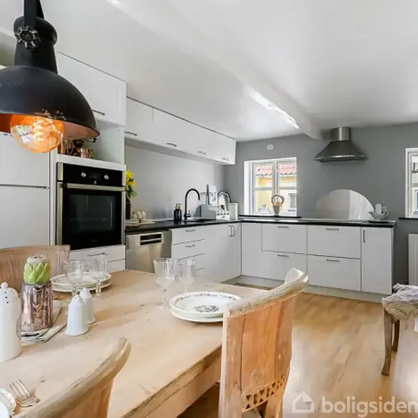 Et moderne køkken udstyret med hvide skabe og indbygget ovn. En træspisebord med stole står i forgrunden. Der hænger en stor sort lampe over bordet.