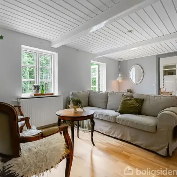 Stue med beige sofa og sofaborde, ved siden af en stol med pels. I baggrunden ses store vinduer med udsigt til træer, en lampe og en spejl på væggen.