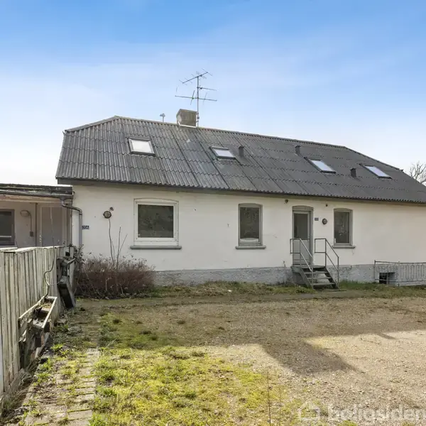 En hvid to-etagers bygning står med skråt tag og flere vinduer. Foran er en grusbelagt gårdsplads omgivet af en lav træhegn. Himmel er klar og solrig.