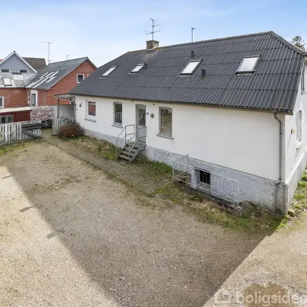 Hvidt hus med sort tag står på en grusvej, omgivet af grønne områder og nærliggende huse. Trappe leder til hovedindgang. Vej ses i baggrunden.