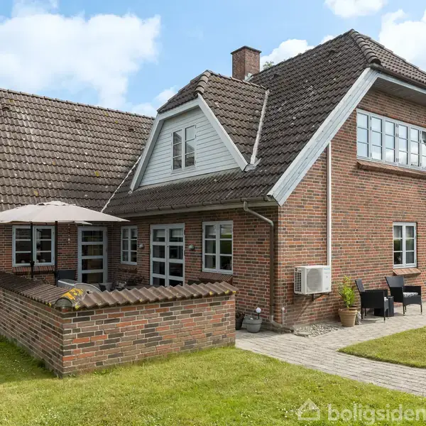 En murstensvilla med en træterrasse og paraply, omkranset af en græsplæne. Huset har store vinduer og et skråt tegltag. En kumulativ atmosfære af et fredeligt boligområde.