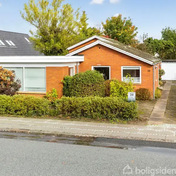 En rød murstensvilla står stille i et villakvarter med en have foran og en dobbeltgarage til højre. Buske omkranser huset, hvor nummeret "30" er synligt.