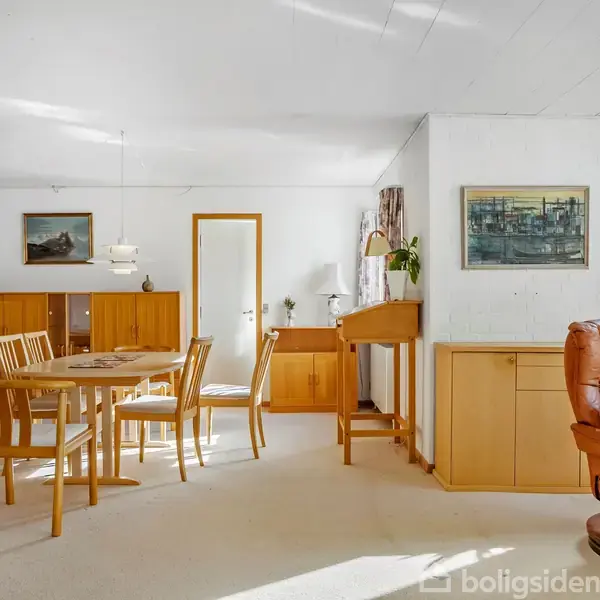 Et møbleret spisestueområde med et træbord og stole omgivet af skabe og kunst på væggene. Store vinduer giver naturligt lys, og der står en lænestol i rummet.