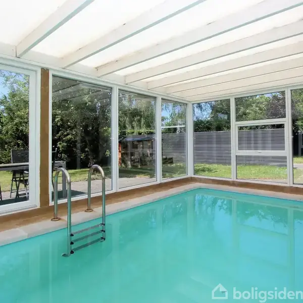 En indendørs swimmingpool med stige, omgivet af store vinduer, der giver udsigt til en grøn have. Sollys strømmer ind gennem et gennemsigtigt tag.