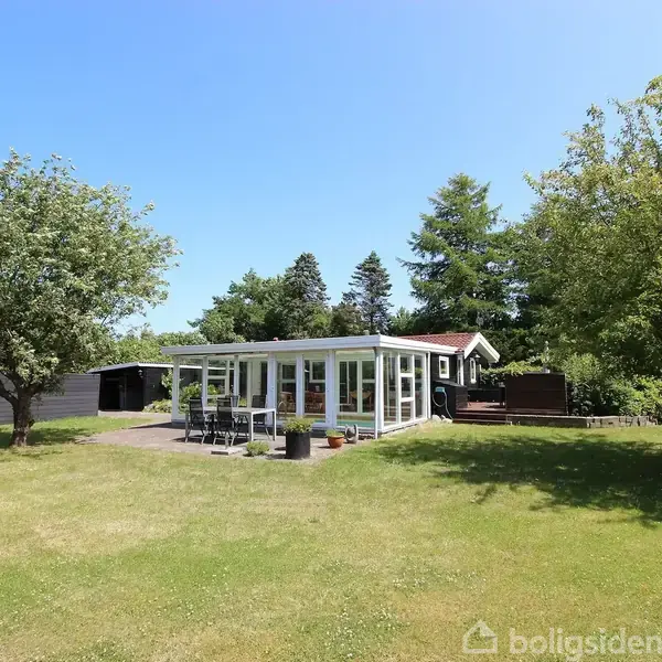 Et lyst hus med store vinduer står stille i en grøn have omkranset af træer og en blå himmel.