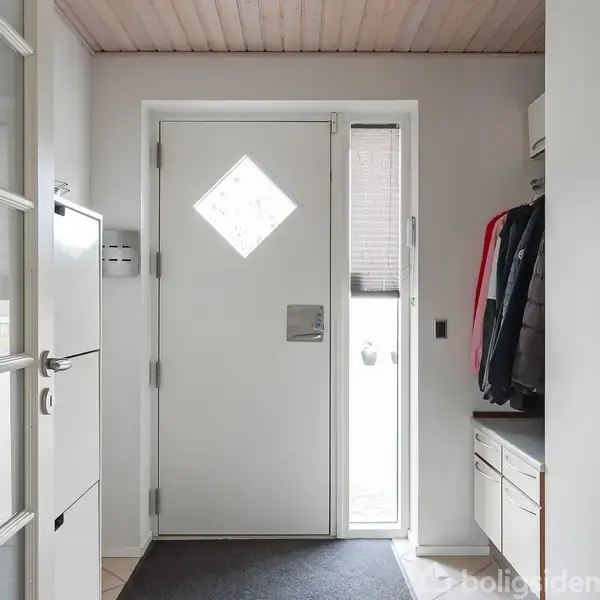 Hoveddør med diamantformet vindue, omgivet af hvid væg og træloft. Til højre hænger jakker ved siden af en kommode; til venstre ses en dør med glasruder.