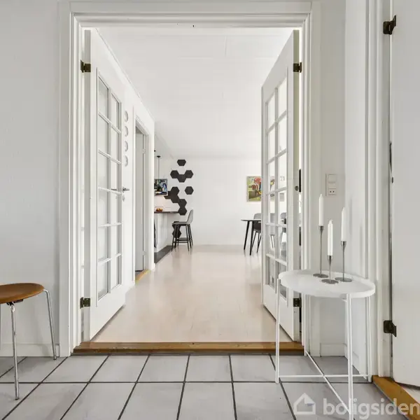 Dobbelte glasdøre åbner til et lyst rum. Fliser på gulvet og lyse trægulve er synlige. Rundt bord og stole står i rummet. Vægkunst hænger i baggrunden.