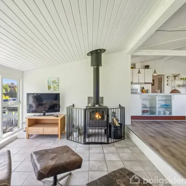Brændeovn midt i stue opvarmer rummet ved at brænde træ. Omkring er der lys stue med sofa, tv, store vinduer og udsigt til terrasse og have.