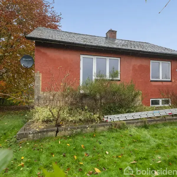 Rødt murstenshus med sort tag, omgivet af en frodig have med græs og træer. En hvid stige ligger ved husets fundament. Skiltning viser "boligsiden" nederst.