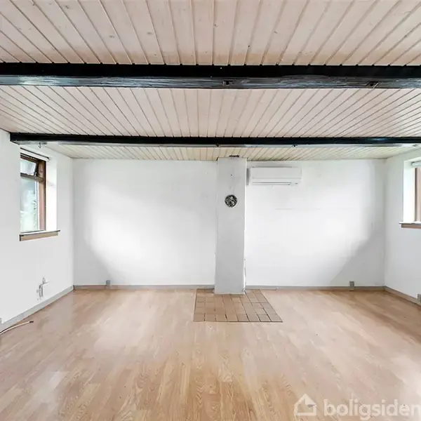 Et tomt rum med trægulve og hvidmalede vægge. Midt i rummet står en hvid søjle. Vinduet på hver side lukker naturligt lys ind. Ventilationsanlæg monteret på væggen.