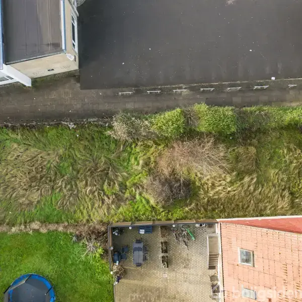 Græsplæne med ujævn bevoksning flankeres af bygninger og gårdhave; kontekst omfatter tag, asfaltvej og en trampolin i haven.