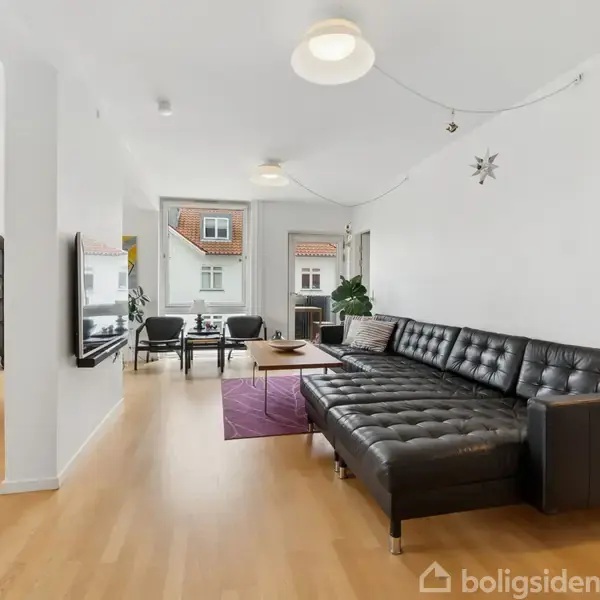 Sort lædersofa står ved en hvid væg. Sofaen vender mod et opholdsområde med et fyrretræsbord. Baggrunden viser store vinduer, der bringer lys ind i rummet.