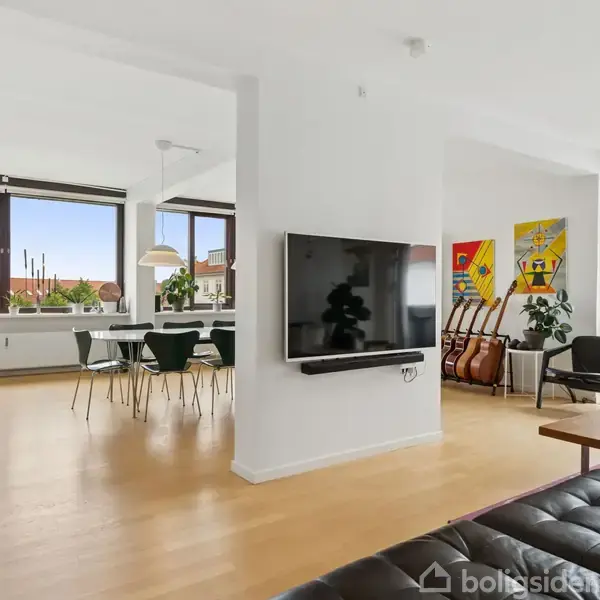 Et moderne stueområde med en fladskærms-TV monteret på en central søjle, omgivet af lyse træmøbler, kunstværker på væggen, store vinduer og spisebord i baggrunden.