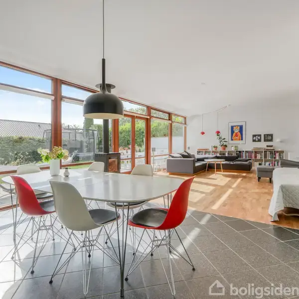 En rund spisebord med stole står i en lys stue. Gulv-til-loft-vinduer slipper masser af lys ind. Stuen har trægulve og moderne møbler med grønne planter udenfor.