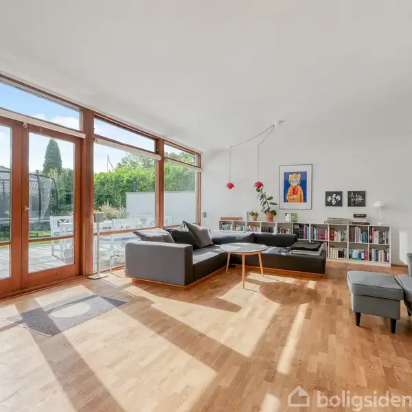 Stue med gråt sofahjørne og sofabord, omkranset af trægulv. Store vinduer åbner op til en have med trampolin. Væggen har en reol, tv og kunstværker.