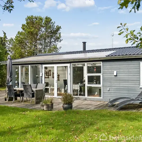 Et gråt sommerhus med store vinduer står stille. Huset har en træterrasse med havemøbler og liggestole. Det er omgivet af en grøn have med træer og buske.