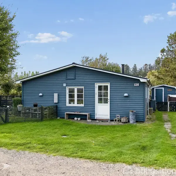 Et blåt hus med en hvid dør står i en grøn have. Stien fører til en terrasse, omgivet af grønne træer og buske. Nummeret "79" er synligt.