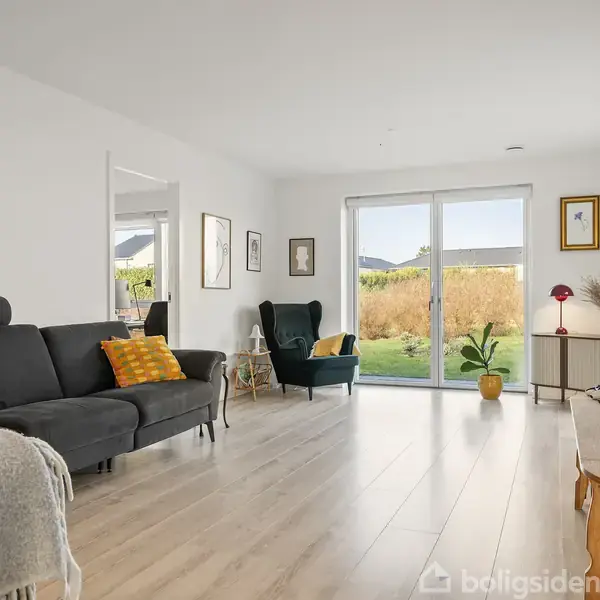 Sort sofa står i stue ved siden af grøn lænestol, med udsigt til have gennem store glasdøre. Lyse trægulve, minimalistisk indretning, og en TV-bænk langs højre væg.