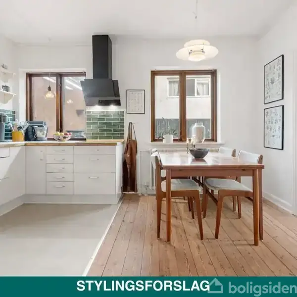 Køkken med hvide skabe og grønne fliser (objekt) står ubrugt (handling) i et lyst rum med træbord og stole nær vinduer; teksten siger "STYLINGSFORSLAG" (kontekst).