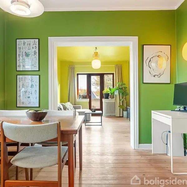Et spisebord med stole står i et rum med lysegrønne vægge, dekoreret med lys og kunst. I baggrunden ses en åbning til en lysere stue med sofa og planter.