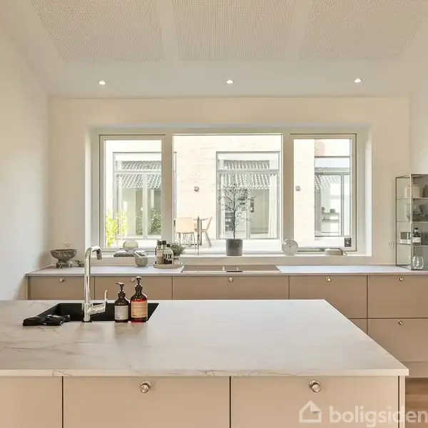 Køkkenbord med vask og sæbedispenser, placeret foran et stort vindue med udsigt til en gårdhave. Bordpladen er lys, og omgivelserne er minimalistiske og moderne.